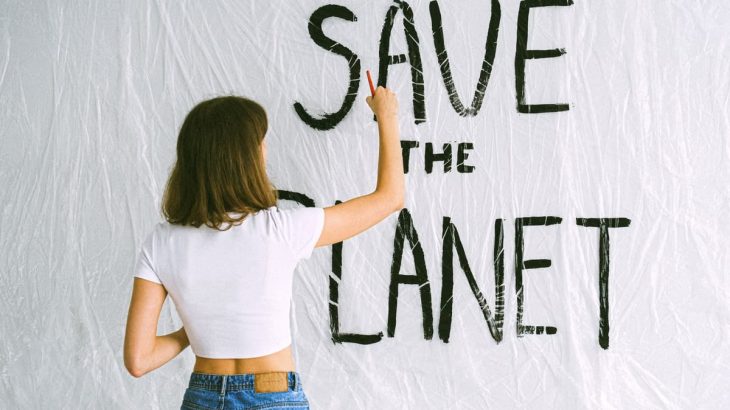 A woman in jeans and white top paints 'Save the Planet' on a wall, advocating environmental awareness.
